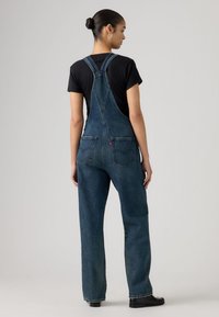 Γυναίκα με τα μαλλιά της σε κότσο, φοράει σκούρα μπλε τζιν σαλοπέτα και μαύρο κοντομάνικο μπλουζάκι, στέκεται γυρισμένη προς τα πίσω μπροστά σε απλό γκρι φόντο.