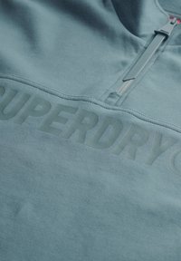 Lichtblauwe fleece pullover met een halve rits en een reliëf "SUPERDRY" logo op de voorkant. Zachte textuur, casual ontwerp, minimale details.