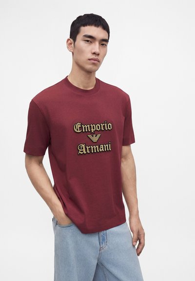 Млад мъж с къса черна коса носи бордо тениска Emporio Armani и светло сини дънки, стои пред обикновен бял фон.