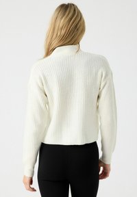 Suéter blanco de punto con un diseño corto, mangas largas y textura acanalada. La parte posterior cuenta con un cuello alto y detalles de costura mínimos.