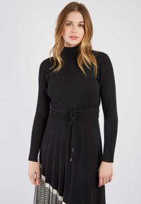 Schwarzes gestricktes Kleid mit einem gerippten hohen Kragen, taillierter Oberseite und einem plisseeartigen Rock mit einem Hahnentritt-Muster am Saum. Mit Kordelzug in der Taille.