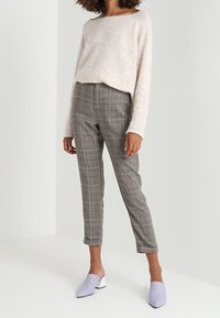 Pull en beige clair avec un col large, associé à un pantalon à carreaux gris avec des accents rouges. Des mules lavande complètent le look avec un design élégant.