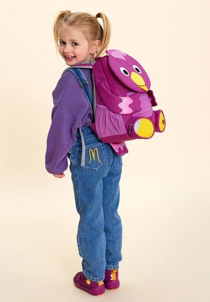 Lächelndes junges Mädchen mit blonden Zöpfen, das einen lila Pullover, blaue Jeans mit gelbem "M", lila Schuhe und einen rosa rucksack in Vogel-Form trägt.