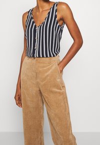 Personne portant un haut sans manches bleu marine et blanc à rayures verticales, boutonné et rentré dans un pantalon large en corduroy tan taille haute.