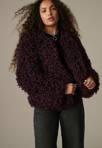 Lilla fluffy jakke med et tekstureret, krøllet stof. Har brede ærmer og en afslappet pasform, parret med mørke jeans og lagdelte ringe.