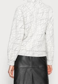 Valkoinen tikattu takki, jossa harmaiden viivojen pintakuvio. Takissa on korkea kaulus ja resorit hihansuissa, yhdistettynä mustaan nahkahameeseen.