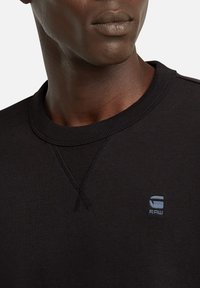 Schwarzer Sweatshirt mit Rundhalsausschnitt, verziert mit einem gekreuzten Stichmuster und einem kleinen blauen "G-RAW" Logo auf der Brust. Weicher Stoff.