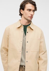 Μπεζ ελαφρύ βαμβακερό μπουφάν με κουμπιά τύπου snap, δύο εμπρόσθιες τσέπες και διακριτικό κόκκινο λογότυπο στο στήθος. Φοράται πάνω από πράσινο πουκάμισο και μπεζ πλεκτό.