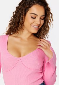 Top lungo a maniche con costine color rosa, scollatura profonda a cuore e cuciture curve. Presenta maniche aderenti con bordi grezzi per un effetto di texture.