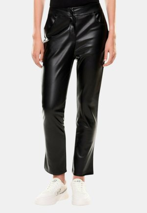 Persona che indossa pantaloni neri in ecopelle e sneaker bianche EA7, in piedi contro uno sfondo grigio chiaro uniforme.