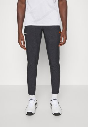 Muž v úzkých černých teplácích s malými logy u kapes, bílých ponožkách, bílých a černých sportovních botách a bílém tričku.