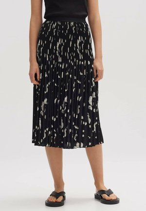 Frau trägt einen schwarzen Midirock mit weißen abstrakten Flecken, kombiniert mit einem schwarzen ärmellosen Oberteil und schwarzen Plateau-Sandalen.