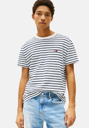 Gestreept wit en marineblauw t-shirt met een ronde hals en korte mouwen. Heeft een klein merkembleem op de borst, vergezeld door lichtblauwe spijkerbroek.
