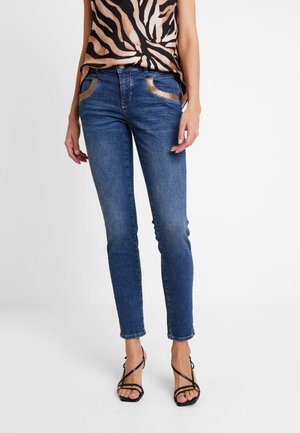 Frau trägt blaue Slim-Fit-Jeans mit metallischen Details an den Taschen, ein Oberteil mit Tiermuster und schwarze Riemchensandalen, steht vor weißem Hintergrund.