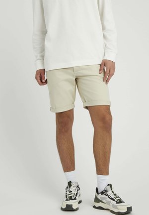 Shorts en coton beige avec des ourlets retournés, associés à une chemise blanche à manches longues et des baskets multicolores avec une semelle texturée et des lacets.