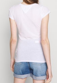 White short-sleeved shirt made from lightweight fabric, featuring a rounded neckline and a slightly longer back hem. Paired with denim shorts.