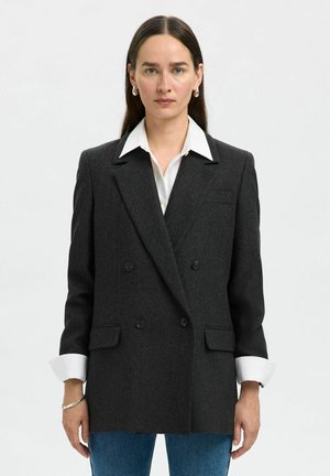 Blazer noir à double boutonnage en gris foncé avec texture chevrons, revers crantés et deux poches avant. Porté sur une chemise blanche à col.