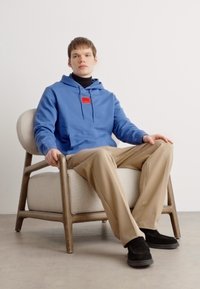 Blå hettegenser med en rød detaljlabel, svart genser, beige bukser og svarte sko. Modell sittende i en lysfarget polstret stol.