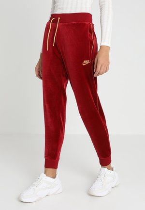 Osoba w czerwonych welurowych spodniach dresowych Nike z żółtym logo i sznurkiem, białym prążkowanym topie i białych sneakersach Nike, stojąca w pomieszczeniu.
