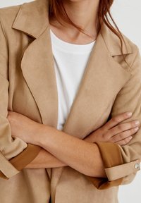 Personne aux longs cheveux portant une veste beige par-dessus une chemise blanche, les bras croisés sur la poitrine, les manches retroussées avec le détail des boutons visible.