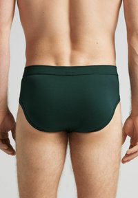Ciemnozielone kąpielówki wykonane z gładkiego materiału, charakteryzujące się niskim stanem i elastycznym pasem, z dopasowanym krojem i minimalnymi szwami.