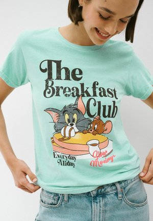 Junge Person trägt ein mintgrünes T-Shirt mit den Cartoonfiguren Tom und Jerry, die über Pfannkuchen mit Sirup hervorschauen, Text „The Breakfast Club“ und „Slow Morning“.