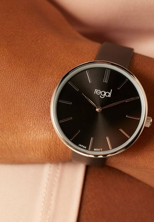 Wristwatch with black minimalist dial, silver case, thin hour markers, and brown leather strap worn on a person's wrist.