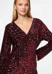 Burgundy paljettklänning med V-ringning och långa ärmar. Har ett texturerat tyg med glänsande, reflekterande paljetter som täcker ytan.
