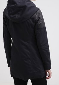 Chaqueta oscura con capucha, que presenta una superficie lisa, mangas de textura contrastante y un corte entallado que se extiende hasta la mitad del muslo.