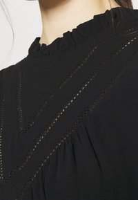 Sort bluse med en bølget høj hals, indviklet udsnit, og blødt tekstureret stof med samlede detaljer langs siderne.