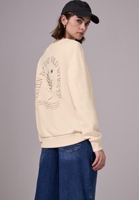 Felpa beige con collo a giro e maniche lunghe, con la scritta "ASCOLTA IL SUSSURRO SELVAGGIO" stampata sul retro. Abbinata a jeans blu a gamba larga.