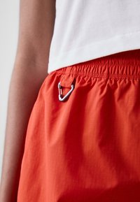 Orange shorts with a drawstring waistband, featuring a silver metal carabiner clip on the front. The fabric has a subtle grid pattern.