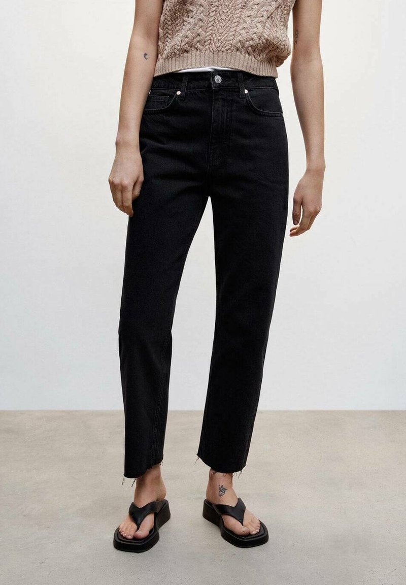 Personne portant un jean noir taille haute avec des ourlets bruts, un top beige en maille sans manches, des sandales à plateforme noires et un petit tatouage à la cheville.