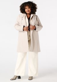 Beige wollen blend jas met een capuchon, met een open voorkant, trekkoorden en zijzakken. Gecombineerd met crèmekleurige broek met wijde pijpen en gebatikte laarzen.