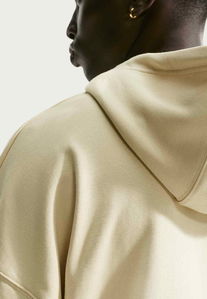 Personne montrée de profil, portant un hoodie beige clair et une petite boucle d'oreille en or, sur un fond neutre.