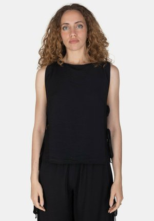 Donna con capelli ricci che indossa un top nero senza maniche e pantaloni neri, in piedi davanti a uno sfondo chiaro e uniforme.