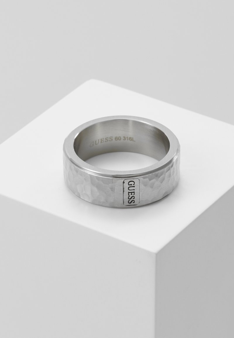 Bague en métal martelé ton argent sur un socle blanc avec une étiquette de marque "GUESS" sur la surface extérieure et "GUESS 60 316L" gravé à l'intérieur.