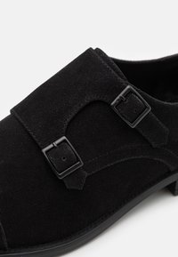 Chaussure en daim noire avec deux boucles noires réglables, un bout arrondi et une finition texturée avec des détails de couture visibles sur le côté.