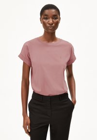 Kurzärmliges T-Shirt aus sanftem pinkfarbenem Baumwollstoff mit umgeschlagenen Ärmeln und subtiler Logodetailierung auf der Brust. Kombiniert mit schwarzen Hosen.