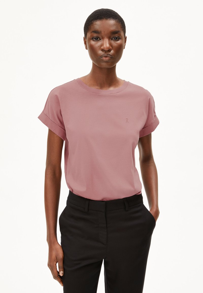 Kurzärmliges T-Shirt aus sanftem pinkfarbenem Baumwollstoff mit umgeschlagenen Ärmeln und subtiler Logodetailierung auf der Brust. Kombiniert mit schwarzen Hosen.