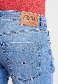 Pantalones vaqueros de denim azul claro con una etiqueta de cuero marrón de Tommy Jeans. Cuenta con un bolsillo trasero con una pequeña etiqueta de logo. Textura suave, corte clásico.