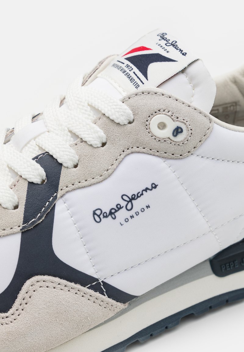 Zapatilla con una parte superior de tela blanca y acentos de ante gris. Presenta un diseño de swoosh azul marino, cordones blancos y una suela de goma negra.