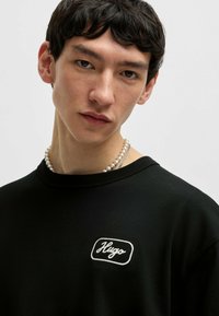 Zwarte sweatshirt met een witte geborduurde "Hugo" logo in een rechthoekige patch. Model draagt een parelketting. Eenvoudig, minimalistisch design.