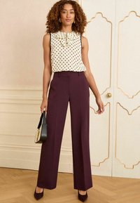 Débardeur crème sans manches avec des pois noirs et un détail en noeud, associé à un pantalon prune taille haute et pantalon large, et des talons assortis. Petit sac à main noir.