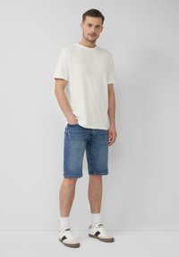 Witte t-shirt met korte mouwen en ronde hals, gecombineerd met blauwe spijkerbroekshorts. Sneakers met witte en beige accenten; witte sokken zichtbaar.