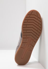 Chaussure en daim gris avec semelle extérieure en caoutchouc brun, présentant un motif de bande de roulement texturé et un logo. Le panneau latéral affiche un contraste de matériau lisse.