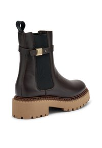 Bota de cuero marrón hasta el tobillo con una robusta suela de goma beige, paneles laterales elásticos y una correa decorativa con un acento en tono dorado.