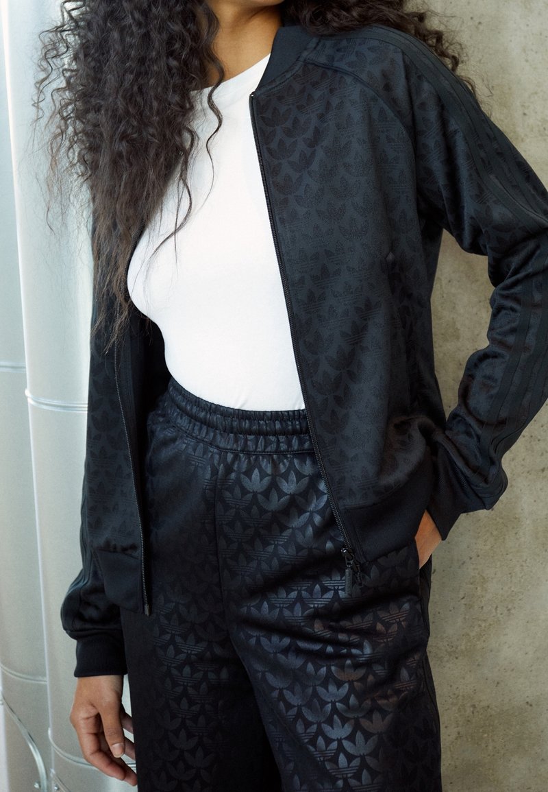 Svart idrottsjacka med subtil mönstrad textur, dragkedja och ribbade manschetter, i kombination med matchande byxor med samma design.