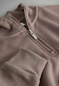 Mikina na zip v měkké, tlumené růžové tkanině. Obsahuje vysoký límec, stříbrný zip a žebrované manžety s texturovaným detailem na látce.