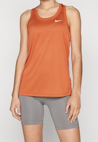 Vrouw in een oranje Nike mouwloos sporttop en grijze strakke fietsshorts, staande tegen een effen achtergrond.